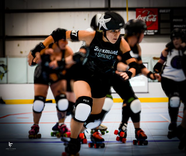 Roller derby skaters lean into a turn during a bout, led by a skater in a black jersey.
